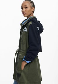 Olivgrüne Jacke mit marineblauem Kapuzen, tailliertem Schnitt und frontalen Taschen. Verziert mit gestickten Details auf Brust und Ärmeln, aus verschiedenen Materialien.