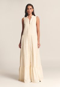 Dolga obleka v kremni barvi z gumbi spredaj, brez rokavov, večstopenjska krila in teksturiranim vzorcem. Mehka tkanina z nežnimi detajli.