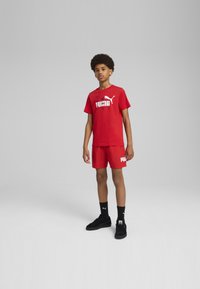 Camiseta de algodón roja con el logo blanco de "PUMA", combinada con pantalones cortos rojos que presentan el mismo logo. Zapatillas negras y calcetas complementan el conjunto.