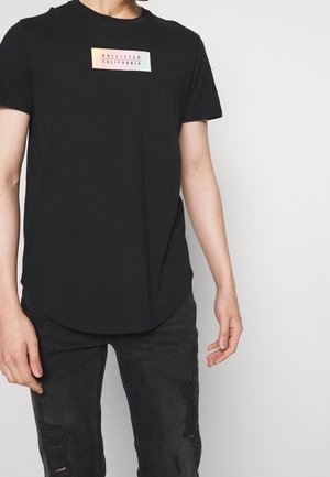 T-shirt noir avec un ourlet arrondi, arborant un écusson logo vibrant indiquant "HOLLISTER CALIFORNIA". Confectionné en tissu doux et léger.