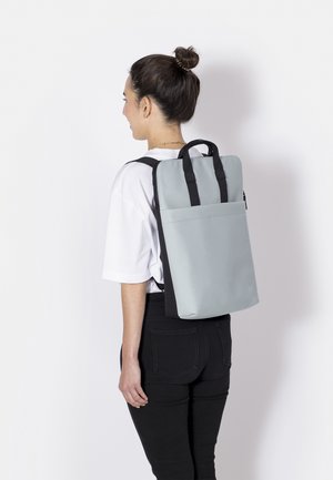 Frau mit braunem Haar in einem Dutt, trägt ein weißes Hemd und schwarze Hosen, trägt einen hellgrauen rechteckigen Rucksack mit schwarzen Trägern.