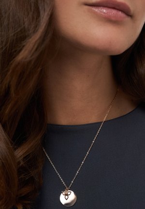 Roségoldene Halskette mit einem runden Anhänger, der ein filigranes Design zeigt, liegt auf einem dunkelblauen Oberteil, mit langem, lockerem braunem Haar, das sichtbar ist.