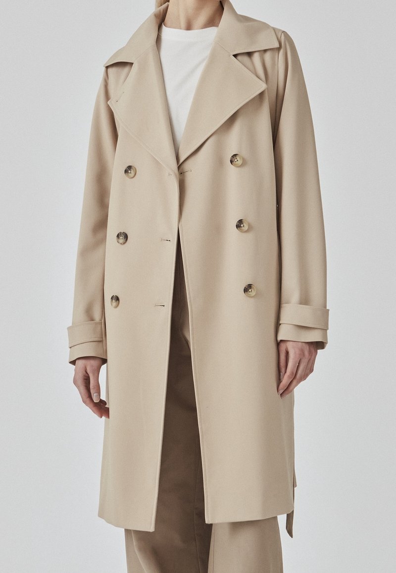 Beige dubbelgeknoopte trenchcoat gemaakt van gladde stof. Heeft een brede kraag, grote knopen en omgeslagen manchetten met een rechte snit.