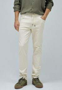 Pantalones beiges con cintura ajustable, bolsillos laterales y un ajuste relajado, combinados con zapatillas de ante verde oliva.