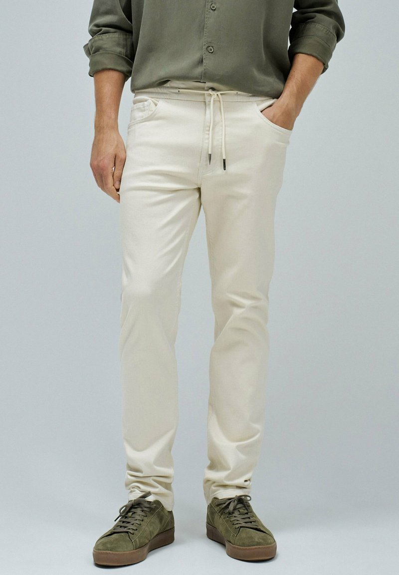 Pantalones beiges con cintura ajustable, bolsillos laterales y un ajuste relajado, combinados con zapatillas de ante verde oliva.