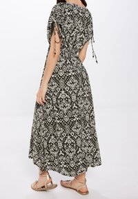 Maxikleid in Grün und Weiß, mit einem gemusterten Design, lockerer Passform und verstellbaren Bindebändern an den Schultern. Getragen mit flachen Sandalen.