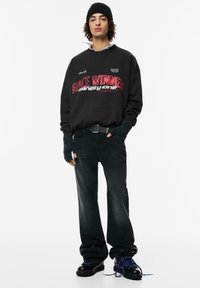 Černá mikina s nápisem "VÍTĚZ ZÁVODU" v červené barvě, černé denimové kalhoty, rukavice bez prstů a černé boty s modrými tkaničkami; uvolněný střih.