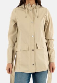 Veste de pluie beige avec devant à boutons, capuche avec cordon de serrage, deux poches avant et texture lisse et imperméable. Conçue pour la fonctionnalité.
