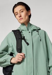 Veste imperméable vert clair avec un col haut, des poignets élastiques et une capuche avec cordon de serrage. Le modèle porte un sac à dos noir.