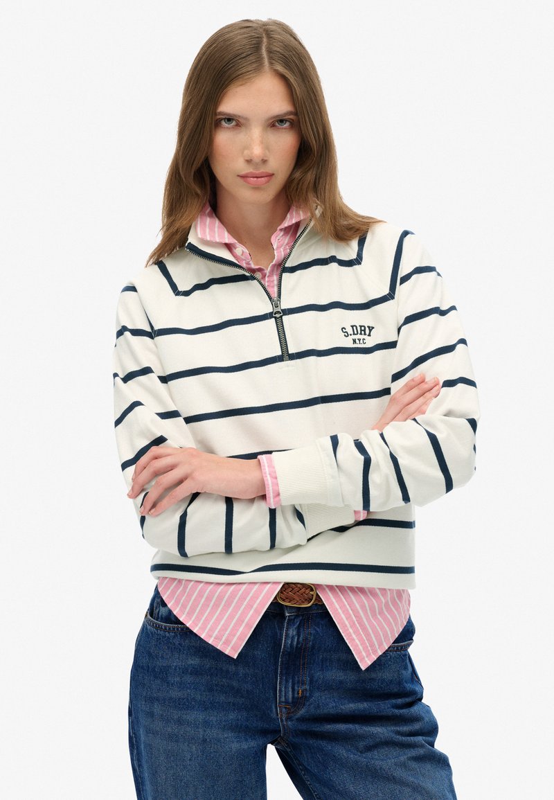 Witte half-zip sweatshirt met marineblauwe strepen, met een logo, gelaagd over een roze gestreept shirt, gecombineerd met blauwe denim jeans.