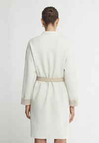 Hvid kjole med bælte, med lange ærmer og beige detaljer ved ærmeåbningerne. Stoffet ser glat og struktureret ud.