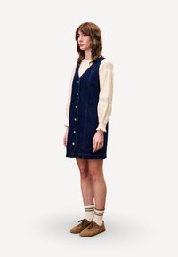 Robe en denim avec un décolleté en V, des boutons sur le devant et des poches latérales. Couleur bleue, coupe droite, superposée à un chemisier crème avec des détails en dentelle.