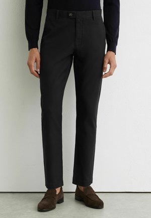 Mann trägt schwarze, eng anliegende Anzughose kombiniert mit dunkelbraunen Wildleder-Loafers, steht vor einer weißen strukturierten Wand.