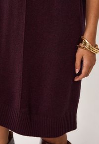 Abito in maglia di un profondo bordeaux con orlo a costine. Presenta una consistenza morbida; abbinato a diversi bracciali d'oro su una mano.