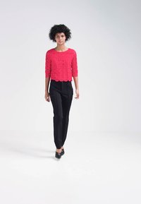 Top corto de encaje rojo con mangas tres cuartos, combinado con pantalones rectos negros y tacones negros, sobre un fondo claro y liso.