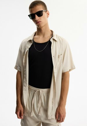 Joven con camiseta sin mangas negra, camisa beige de manga corta con botones, pantalones beige con cordón, collar de cadena, gafas de sol y pendiente, de pie.