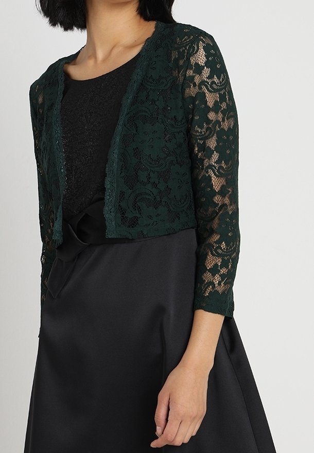 Žena má na sobě tmavě zelený květovaný krajkový krátký kardigan přes černé šaty s atlasovou mašlí v pase, stojí před světlým pozadím.