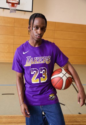Violet medvilninis krepšinio marškinėliai su "LAKERS" ir "23" geltonos ir baltos spalvos. Derinami su mėlynomis džinsomis, laikant krepšinį.