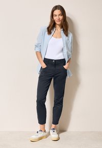 Světle modrá pruhovaná košile na knoflíky, bílý top bez rukávů, námořnické slim-fit kalhoty a bílé tenisky s béžovými podrážkami. Model stojí s rukama v kapsách.