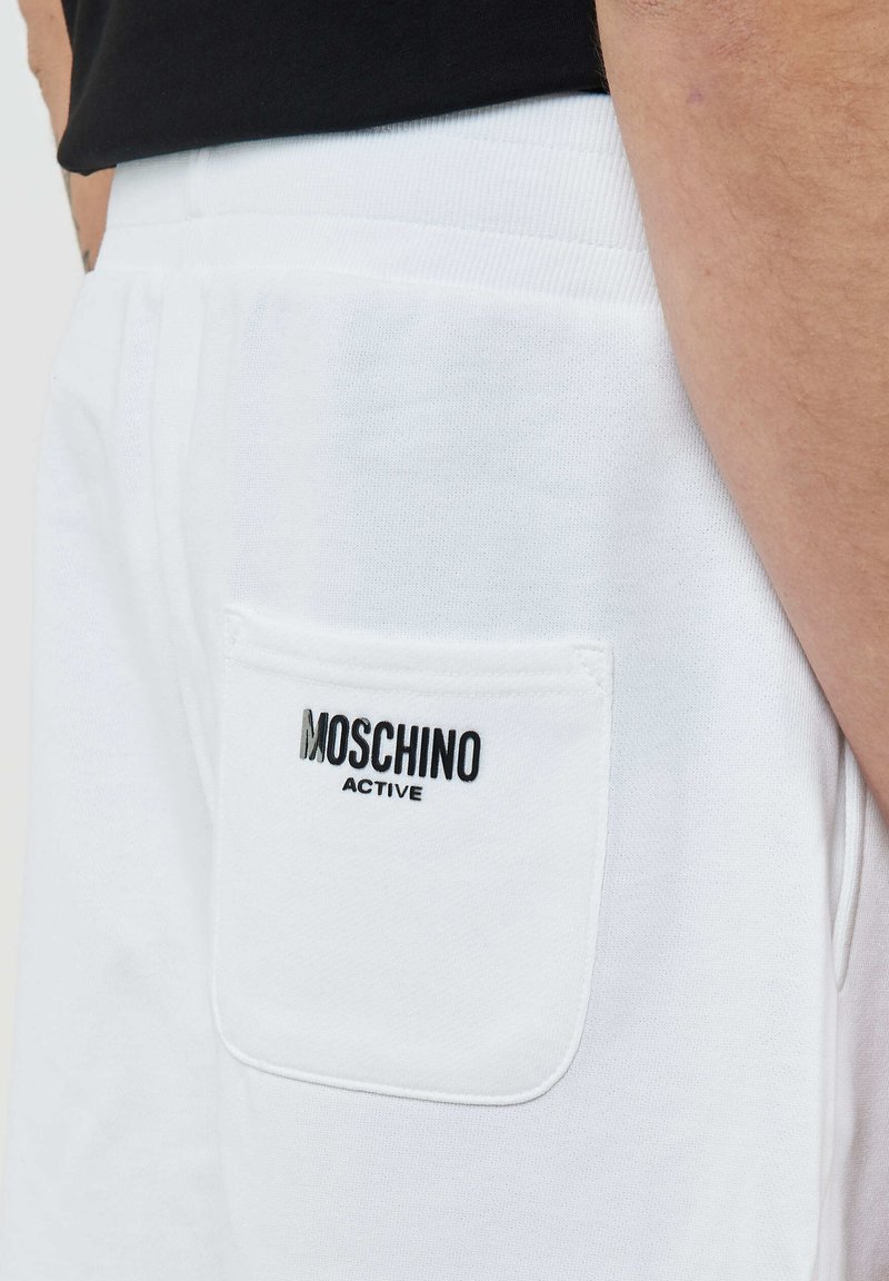 Gros plan sur la poche d'un pantalon de survêtement blanc avec le logo noir "MOSCHINO ACTIVE", porté avec un t-shirt noir, montrant une partie du bras du porteur.