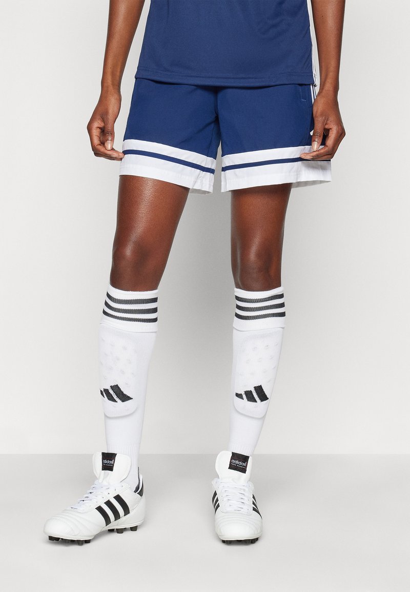 Pantalones cortos deportivos azul marino con rayas blancas, combinados con calcetines blancos con acentos negros y botas negras.