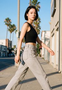 Giovane donna con capelli corti neri che indossa un crop top nero e jeans grigio chiaro mentre cammina su un marciapiede soleggiato fiancheggiato da palme.