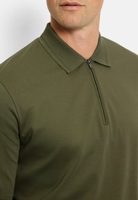 Olivgrünes Polo-Shirt mit einem halben Reißverschluss am Kragen, aus glattem Stoff gefertigt. Verfügt über einen klassischen Kragen und eine taillierte Passform.