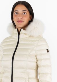 Chaqueta de plumas clara con diseño de acolchado horizontal, que cuenta con una cremallera negra y un ribete de pelo sintético en la capucha. Parche negro con logo en la manga.