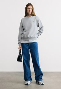 Femme portant un sweat-shirt gris, un jean large bleu, des baskets blanches, tenant un sac à main noir, debout devant un fond clair uni.