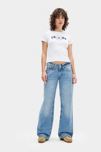 Kvinne med krøllete hår iført en hvit croppet t-skjorte med teksten "1998", løse lyseblå jeans og brune mokkassinsko, stående mot hvit bakgrunn.