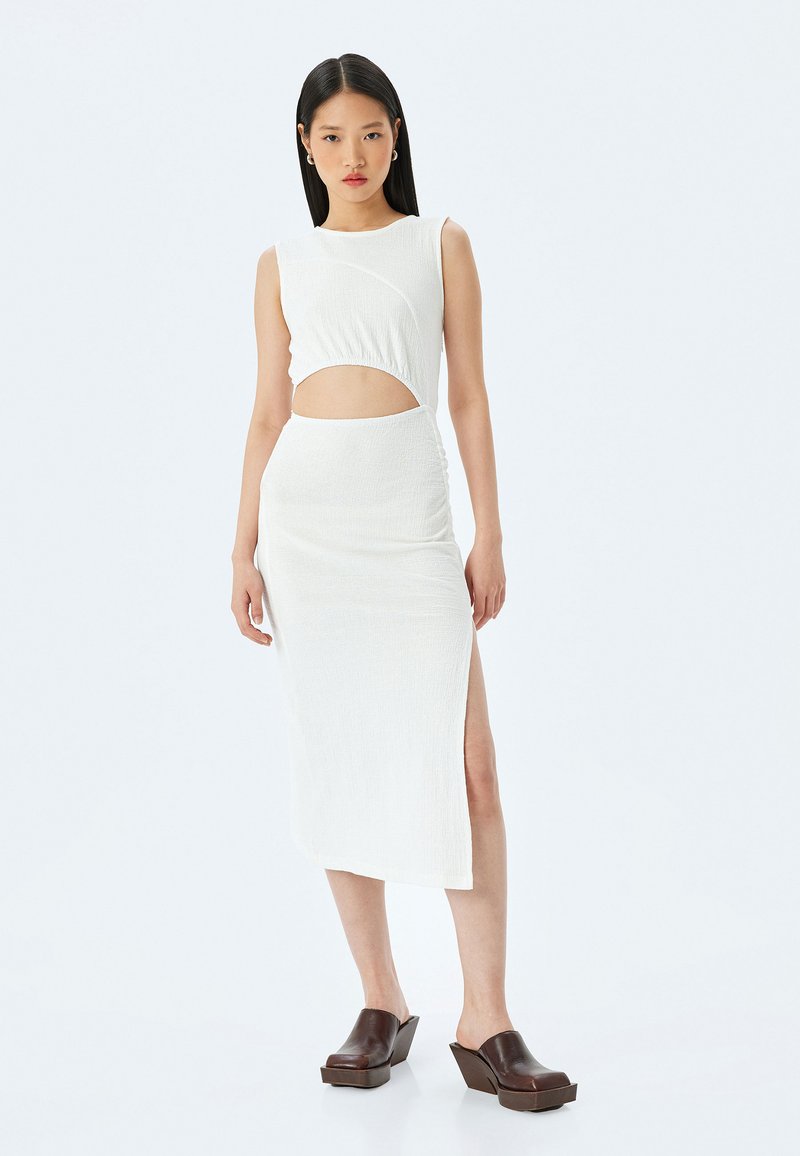 Witte tweedelige outfit met een mouwloze top met uitsnijding, een aansluitende midi-rok met een zijsplit en een textuurstof. Bruine wedge klompen.