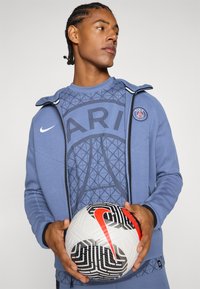 Felpa sportiva blu con un motivo geometrico e testo "PARIGI", abbinata a una giacca con zip coordinata; tiene in mano un pallone da calcio decorato in bianco e nero.