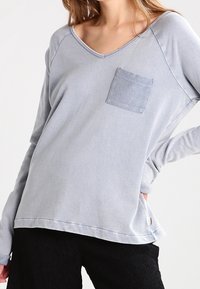 Maglietta a maniche lunghe blu chiaro con scollo a V, dotata di una piccola tasca sul petto e una vestibilità relaxed. Tessuto morbido con dettagli a coste all'orlo.