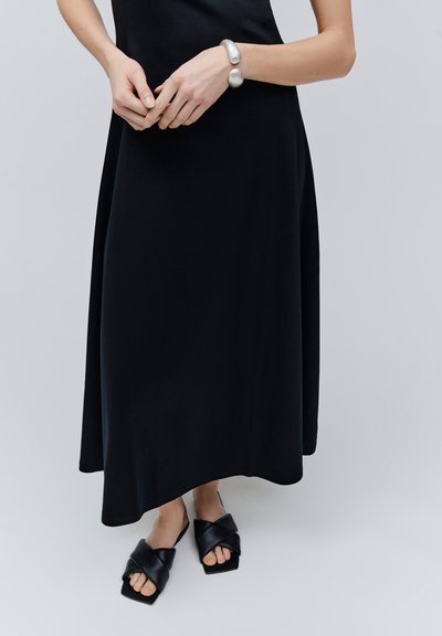 Femme en robe noire sans manches portant un bracelet manchette argenté et des sandales noires à bout carré avec des lanières croisées, debout sur un fond clair.