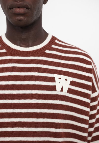 Suéter a rayas en colores óxido y crema, con un cuello redondo y un gran logo en forma de letra "W" blanca en el lado izquierdo. Textura suave.