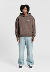 Brown zip-up hoodie with a front logo, paired with light blue loose-fitting jeans and black and white sneakers, standing against a gray background.