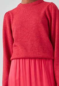 Rød strikket sweater med lange ærmer, struktureret finish, ribbet halsudskæring, parret med en svajende pink nederdel, som er synlig forneden.