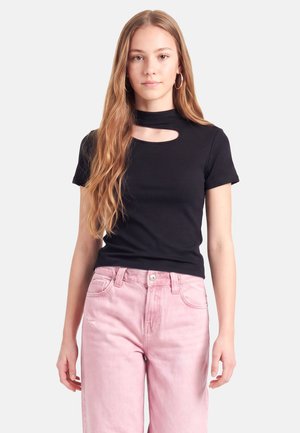 Jeune femme aux longs cheveux ondulés portant un haut noir à manches courtes avec un décolleté découpé et un jean taille haute rose clair.