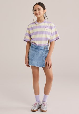 Mädchen steht mit entspannten Händen, trägt ein T-Shirt mit lavendel- und cremefarbenen Streifen, einen Wickelrock aus Denim, lavendelfarbene Socken und silberne Sneaker.