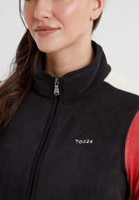Black fleece vest featuring high collar, zip closure, and embroidered "TOG24" logo. Accented with white shoulder panels and red sleeves.