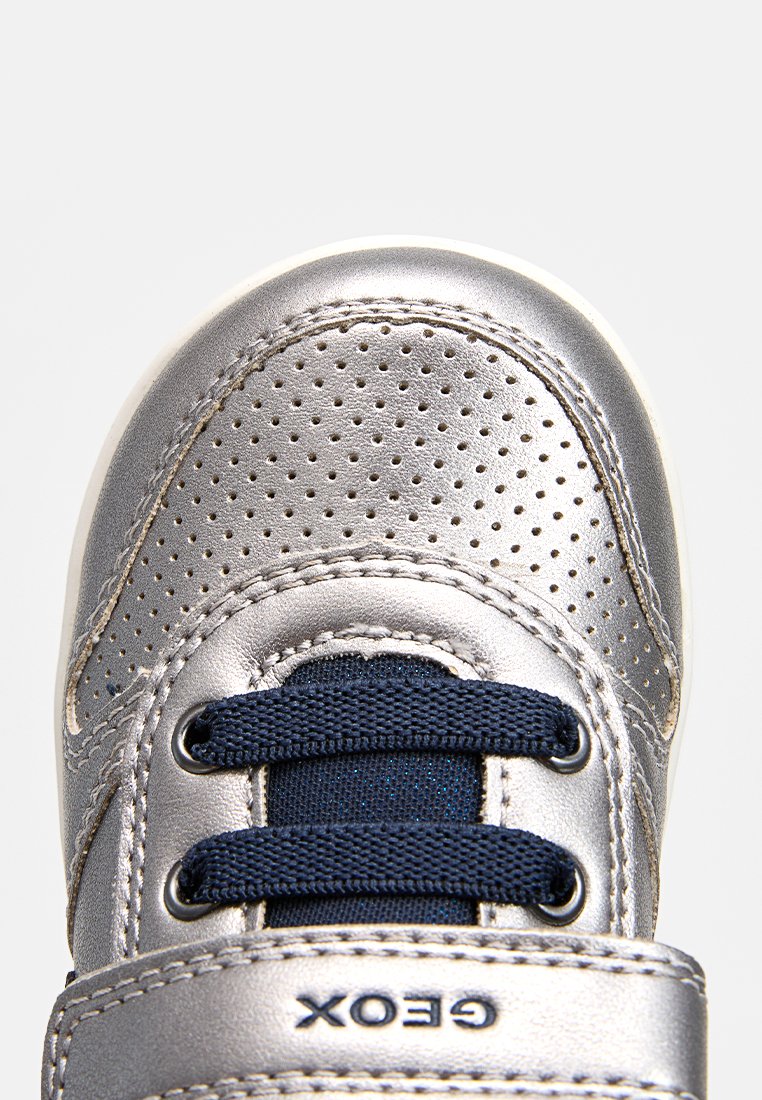 Sneaker argento metallizzato con tomaia perforata, lacci blu navy e strap in velcro. Presenta suola morbida e dettagli cuciti. Logo del brand visibile.