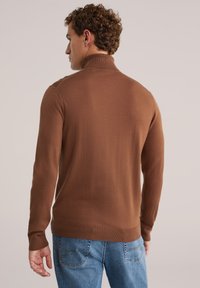 Rjav pulover s turtleneck-ovim izrezom, narejen iz mehkega materiala, z oprijetim krojem in rebrastimi manšeti ter robom, oblečen z svetlo modrimi kavbojkami.