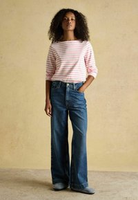 Mujer de pie con una mano en el bolsillo, vistiendo una camisa de manga larga a rayas rosas y blancas, vaqueros anchos de pierna azul y zapatos grises.