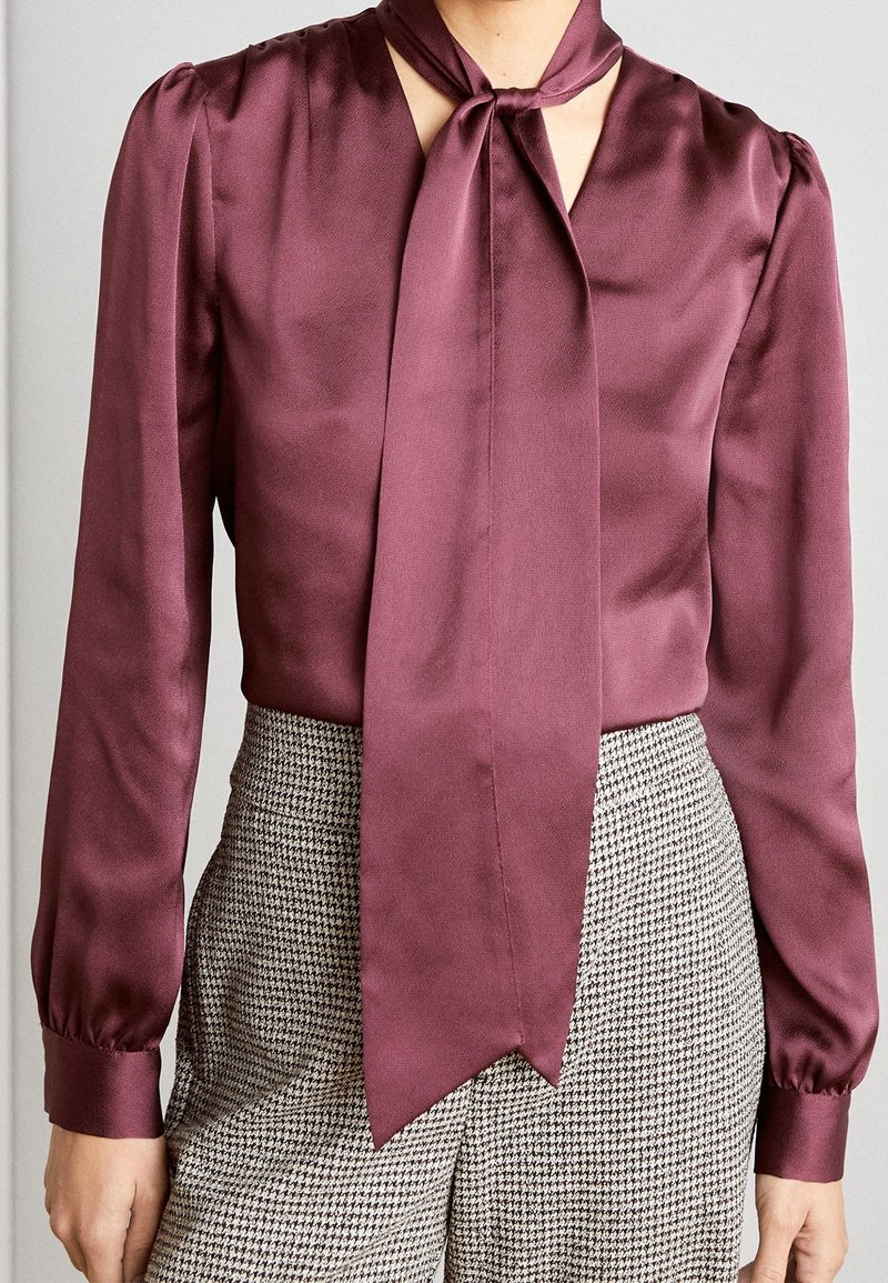 Blouse bordeaux en satin à manches longues avec un foulard noué autour du cou, associée à un pantalon à taille haute noir et blanc motif pied-de-poule.