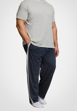 Mężczyzna ubrany w jasnoszarą koszulę z krótkim rękawem i granatowe spodnie z szarymi bocznymi paskami, stoi boso z rękami w kieszeniach.