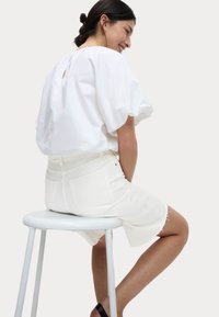 Blusa bianca con maniche a sbuffo, dettaglio a chiave sul retro; abbinata a pantaloni corti bianchi e sandali neri con cinturini, seduta su uno sgabello.