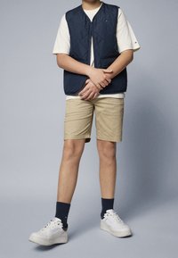 Námořnická prošívaná vesta přes bílém tričku, béžové šortky a bílé tenisky s námořnickými ponožkami. Jednoduchý design a pohodlný střih.