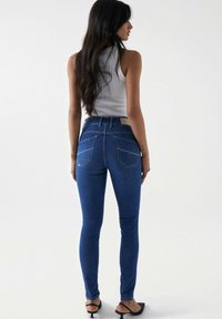 Jeans de mezclilla azul ajustados con una cintura de media altura, que presentan una costura distintiva en los bolsillos traseros y una etiqueta en la cinturilla. Textura suave, diseño ceñido.