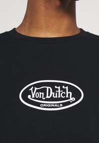 Camiseta de algodón negra con cuello redondo, que presenta un logo blanco que dice "Von Dutch ORIGINALS" en un diseño ovalado.