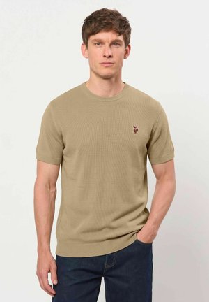 Jeune homme portant un pull en maille beige à manches courtes avec un petit logo brodé, debout avec une main dans la poche devant un fond uni.